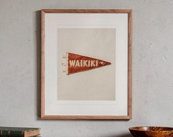 May include: A framed print featuring a triangular red felt pennant with the word WAIKIKI printed in white capital letters and a small white wave graphic on the right side. The pennant has three white fabric ties on the left edge.
