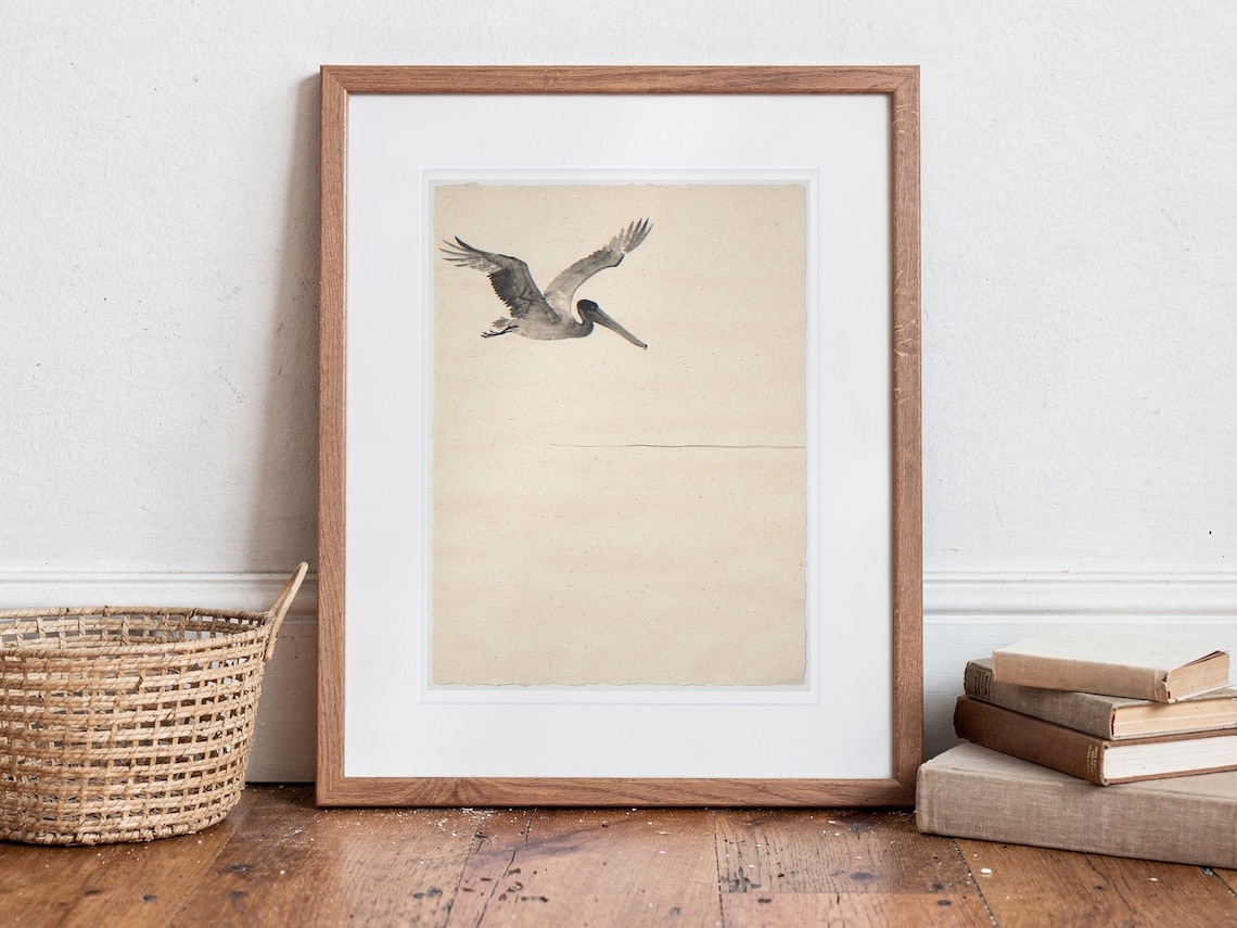 Flying Pelican Ink Style print in a Red Oak frame with a white mat, standing on the floor, leaning back slightly against the wall in a living room floor lean.