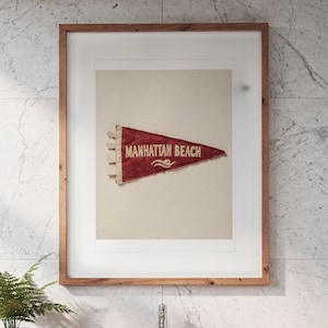 May include: A framed wall art piece featuring a triangular red felt pennant with the text Manhattan Beach printed in white lettering. The pennant includes a small white graphic of a wave and is displayed within a light brown wooden frame.
