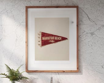 May include: A framed wall art piece featuring a triangular red felt pennant with the text Manhattan Beach printed in white lettering. The pennant includes a small white graphic of a wave and is displayed within a light brown wooden frame.