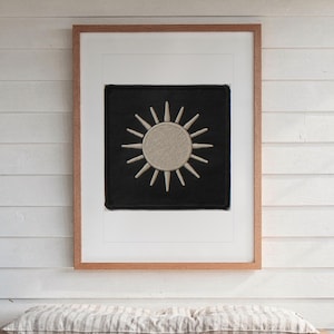 May include: A square black fabric patch featuring an embroidered beige sun with radiating rays, centered within a white mat and a light brown wooden frame. The sun design has a textured circular center and sixteen pointed rays extending outward.