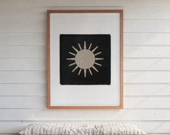 May include: A square black fabric patch featuring an embroidered beige sun with radiating rays, centred within a white mount and a light brown wooden frame. The sun design has a textured circular centre and sixteen pointed rays extending outwards.