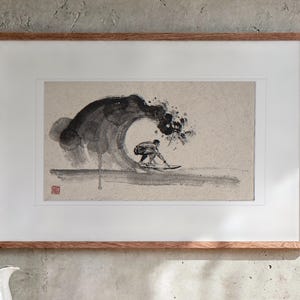 May include: A framed ink wash painting on textured paper depicting a surfer riding inside a large, dark, swirling wave. The artwork features expressive black brushstrokes, a light beige background, and a small red seal stamp in the corner.