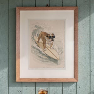 Bulldog Surfing print in a Red Oak frame with a white mat, hanging above the table in a screened beach porch.