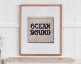 May include: A framed wall art piece featuring a square, cream-colored fabric patch with frayed edges. The text "OCEAN BOUND" is embroidered in dark navy blue thread in a bold, stylized font, centered within the white mat and wood frame.