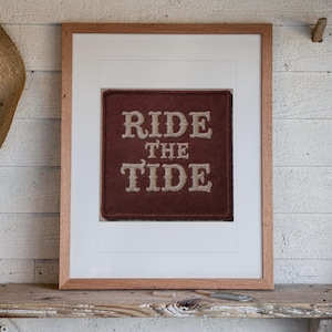 May include: A framed square patch featuring the text RIDE THE TIDE in beige embroidered lettering on a dark brown felt background. The patch is centered within a white mat and enclosed in a light-colored wooden frame with a clean finish.