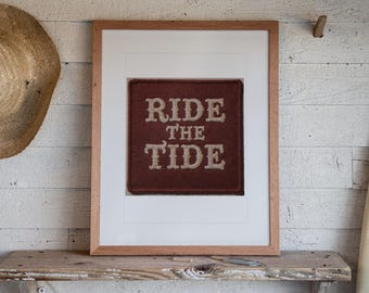 May include: A framed square patch featuring the text RIDE THE TIDE in beige embroidered lettering on a dark brown felt background. The patch is centred within a white mat and enclosed in a light-coloured wooden frame with a clean finish.