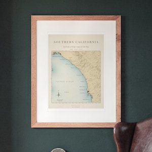 Southern California Surf Map printable art, shown printed in a Red Oak frame with a white mat, hanging on the wall between the shelves in a home library.