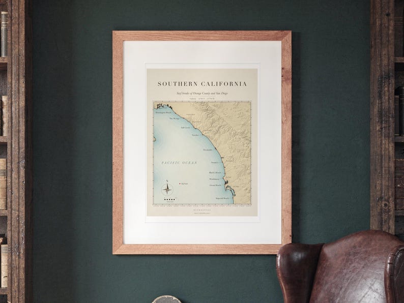 Southern California Surf Map printable art, shown printed in a Red Oak frame with a white mat, hanging on the wall between the shelves in a home library.