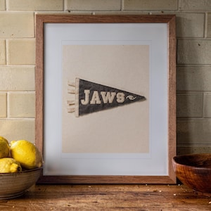 May include: A framed print featuring a dark triangular pennant with the word JAWS and a small wave graphic in white lettering. The artwork is displayed in a simple wooden frame with a white mat, centered against a light-colored background.