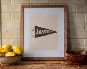 May include: A framed print featuring a dark triangular pennant with the word JAWS and a small wave graphic in white lettering. The artwork is displayed in a simple wooden frame with a white mat, centered against a light-colored background.
