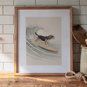 May include: A framed art print featuring a beagle dog wearing sunglasses while standing on a wooden surfboard riding a light green ocean wave. The illustration is set against a neutral beige background and enclosed in a light brown wooden frame.
