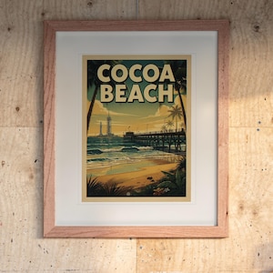 May include: A framed art print featuring a beach scene with the text COCOA BEACH in bold letters. The illustration depicts a wooden pier extending into the ocean, palm trees, and a sunset sky. The print is housed in a light-colored wooden frame.