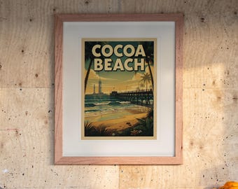 May include: A framed art print featuring a beach scene with the text COCOA BEACH in bold letters. The illustration depicts a wooden pier extending into the ocean, palm trees, and a sunset sky. The print is housed in a light-coloured wooden frame.