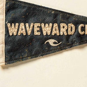 Close detail of the Waveward Club pennant texture, cream lettering, and vintage finish