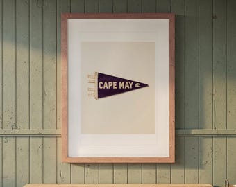 May include: A framed print featuring a dark purple triangular pennant with the text CAPE MAY in white capital letters and a small white wave graphic on the right side, displayed within a light brown wooden frame on a wall.
