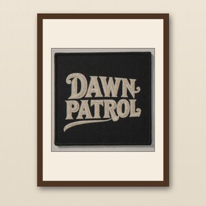Pode incluir: Um patch quadrado preto com as palavras "DAWN PATROL" bordadas em uma fonte creme em estilo vintage. O patch é emoldurado por uma borda marrom e colocado em um fundo neutro.
