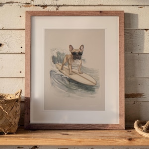 May include: A framed art print featuring a tan French Bulldog wearing black sunglasses while standing on a surfboard riding a wave. The illustration is set against a light beige background and enclosed in a simple brown wooden frame.