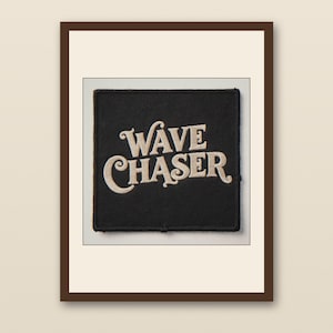 Pode incluir: Um patch quadrado preto com as palavras "WAVE CHASER" bordadas em creme em uma fonte estilizada. O patch é emoldurado por uma borda marrom, sugerindo que pode ser um patch de ferro ou um elemento decorativo para roupas ou acessórios.