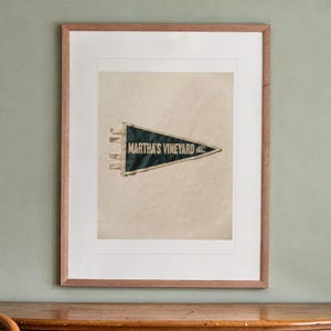 May include: A framed, dark green triangular felt pennant featuring the text "MARTHA'S VINEYARD" in white capital letters, accompanied by a small white wave graphic. The pennant is centered within a white mat and a light brown wooden frame.