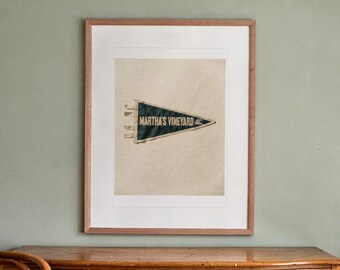 May include: A framed, dark green triangular felt pennant featuring the text "MARTHA'S VINEYARD" in white capital letters, accompanied by a small white wave graphic. The pennant is centered within a white mat and a light brown wooden frame.