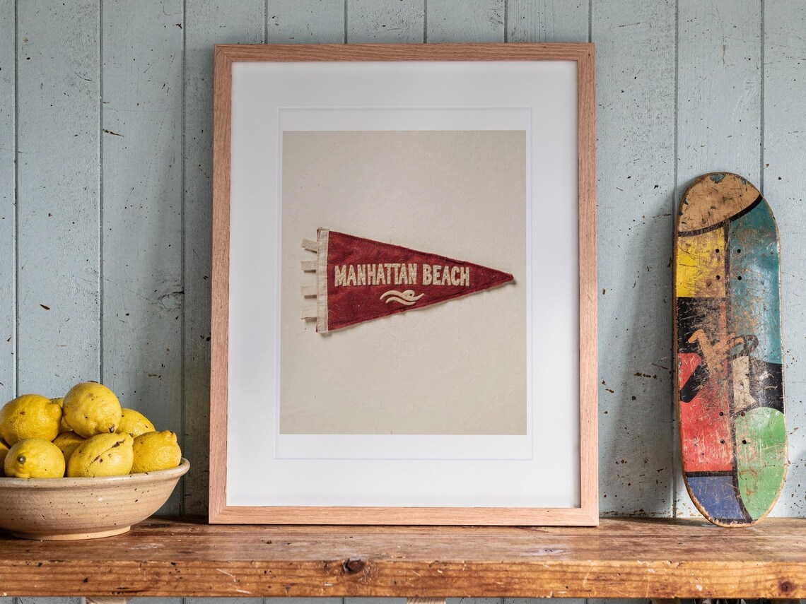 Manhattan Beach Surf Pennant print in a Red Oak frame with a white mat, leaning on the shelf against the wall in a cottage kitchen.