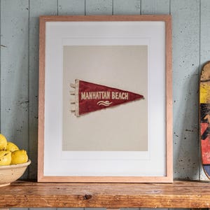 Manhattan Beach Surf Pennant print in a Red Oak frame with a white mat, leaning on the shelf against the wall in a cottage kitchen.