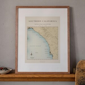 May include: A framed map print titled SOUTHERN CALIFORNIA, Surf Breaks of Orange County and San Diego. The map features a tan landmass and blue Pacific Ocean with a compass rose, all enclosed in a light brown wooden frame with a white mat.