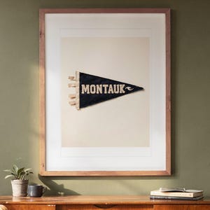 May include: A framed wall art piece featuring a dark navy blue triangular felt pennant centered on a cream background. The pennant displays the word MONTAUK in bold, light-colored capital letters, accompanied by a small wave graphic symbol.
