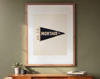 May include: A framed wall art piece featuring a dark navy blue triangular felt pennant centred on a cream background. The pennant displays the word MONTAUK in bold, light-coloured capital letters, accompanied by a small wave graphic symbol.