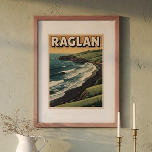 Puede incluir: Un póster de viaje enmarcado con la palabra RAGLAN en letras negras negritas en la parte superior. La ilustración muestra un paisaje costero con colinas verdes, una playa de arena oscura y olas azules bajo un cielo de atardecer.