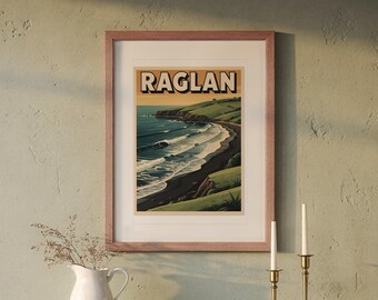 May include: A framed travel poster featuring the word RAGLAN in bold black letters at the top. The illustration depicts a coastal landscape with green rolling hills, a dark sandy beach, and blue ocean waves under a soft sunset sky.