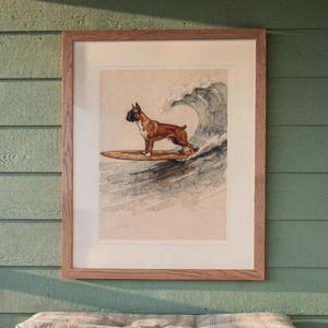Boxer Surfing printable art, shown printed in a Red Oak frame with a white mat, hanging above the bench in a screened beach porch.