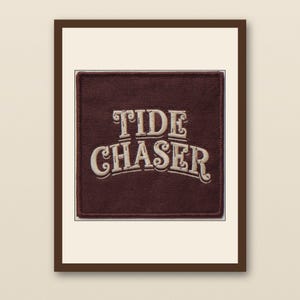 Könnte beinhalten: Ein quadratischer, gerahmter Aufnäher mit dem Text "TIDE CHASER" in einer verzierten, Vintage-Schriftart. Der Text ist cremefarben auf einem dunkelroten Hintergrund. Der Rahmen ist dunkelbraun.
