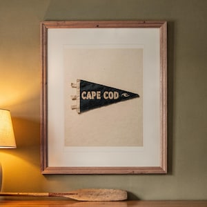 May include: A framed wall art piece featuring a dark blue triangular pennant with the text CAPE COD in white lettering. The pennant is centered on a light cream background within a light-toned wooden frame, displayed on a wall.