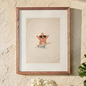May include: A framed wall art print featuring a cartoon starfish wearing black sunglasses while surfing on a surfboard atop a stylized blue wave. The illustration is centered on a textured, light beige background within a wooden frame.