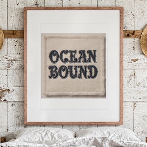 Puede incluir: Un parche de tela cuadrado de color crema con bordes deshilachados que presenta el texto bordado en azul oscuro "OCEAN BOUND" en una fuente estilizada, exhibido dentro de un marco de madera con un borde blanco.