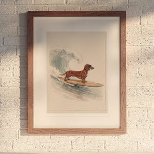 May include: A framed art print featuring a brown dachshund standing on a wooden surfboard while riding a blue watercolor ocean wave. The artwork is enclosed in a simple brown wooden frame with a white mat border, displayed on a white brick wall.