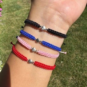 May include: Four friendship bracelets with silver heart charms. The bracelets are red, pink, blue, and black.