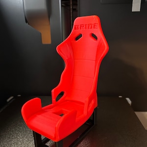 Puede incluir: Un asiento de carreras rojo impreso en 3D con la palabra "BRIDE" impresa en la parte posterior. El asiento está montado sobre una base negra.