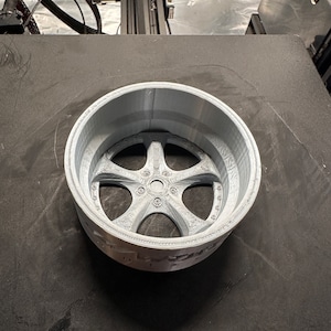 May include: A 3D printed model of a car wheel rim. The rim is silver and has a detailed design with spokes and a centre cap.