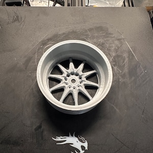 May include: A 3D printed gray car wheel rim with a detailed spoke design. The wheel is sitting on a black surface with the word "Ender" printed on it.