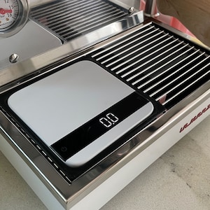 May include: A white espresso machine with a stainless steel drip tray and a digital scale that reads "0.00". The machine has a red "La Marzocco" logo on the front.