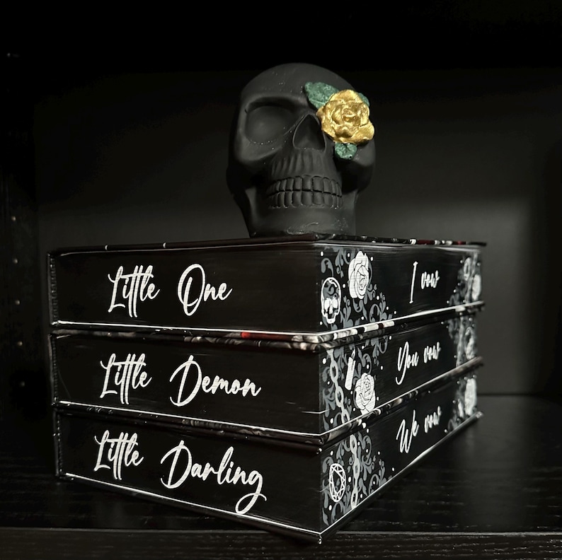 May include: Three black books stacked on top of each other. The books have white text on the front that reads "Little One", "Little Demon", and "Little Darling". Each book also has a black and white design on the front. A black skull with a gold rose is sitting on top of the books.