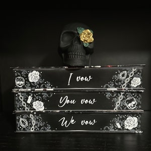 May include: Three black books stacked on top of each other. Each book has a different design on the cover. The top book says "I vow", the middle book says "You vow", and the bottom book says "We vow". A black skull with a gold and green flower on its eye sits on top of the books.