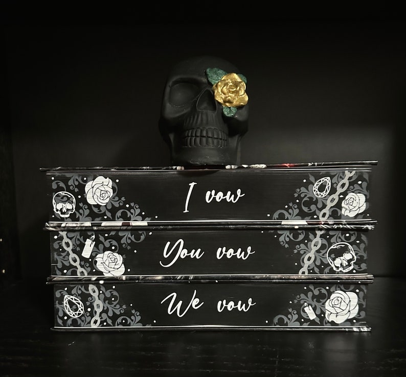 May include: Three black books stacked on top of each other. Each book has a different design on the cover. The top book says "I vow", the middle book says "You vow", and the bottom book says "We vow". A black skull with a gold and green flower on its eye sits on top of the books.