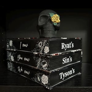 May include: Three black books stacked on top of each other. The books have white text on the front that reads "I vow", "You vow", "We vow". The top book has the name "Ryat's" in white text. The middle book has the name "Sin's" in white text. The bottom book has the name "Tyson's" in white text. A black skull with a gold rose on one eye sits on top of the books.