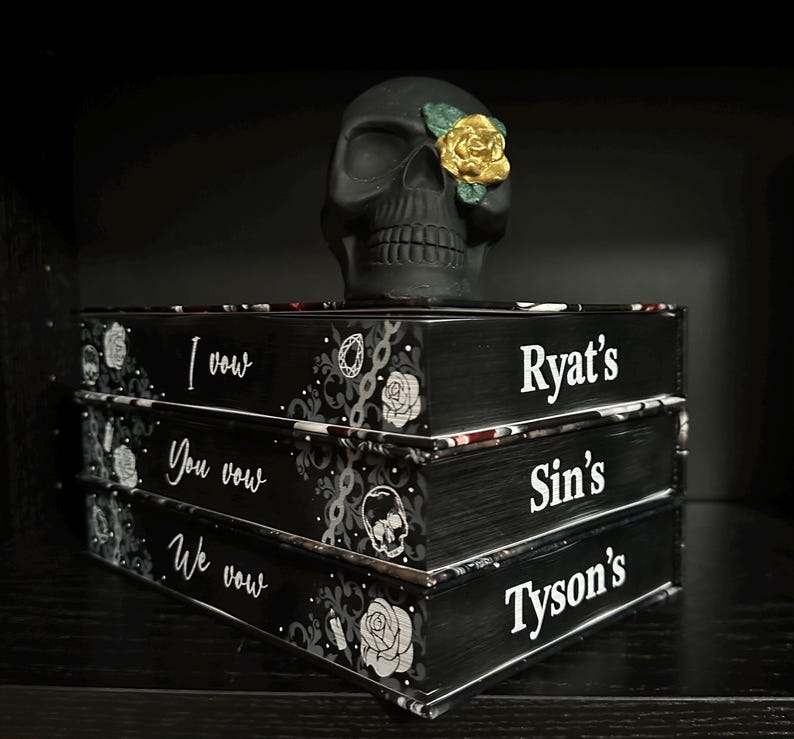 May include: Three black books stacked on top of each other. The books have white text on the front that reads "I vow", "You vow", "We vow". The top book has the name "Ryat's" in white text. The middle book has the name "Sin's" in white text. The bottom book has the name "Tyson's" in white text. A black skull with a gold rose on one eye sits on top of the books.