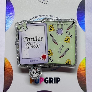 May include: A PopGrip with a glittery border, featuring a book design with the text "Thriller Girlie." The design includes a bow, a skull charm, a button, knives, footprints, and the number "2."
