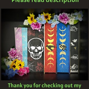 May include: Five decorative books with different designs. The book on the left has a skull and crossbones design. The next book has a moon and stars design. The third book has a snake design. The fourth book has a bird design. The fifth book has a mountain design. The books are decorated with flowers.